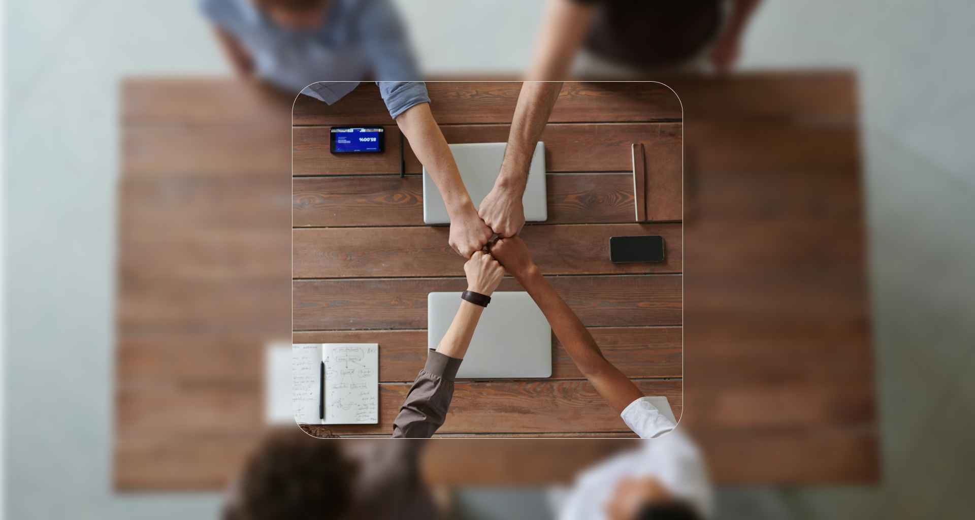 Four people sit around a wooden table, bumping fists in the center. Laptops, a notebook, and phones are on the table. The photo is taken from above, with a highlighted border focusing on their hands.